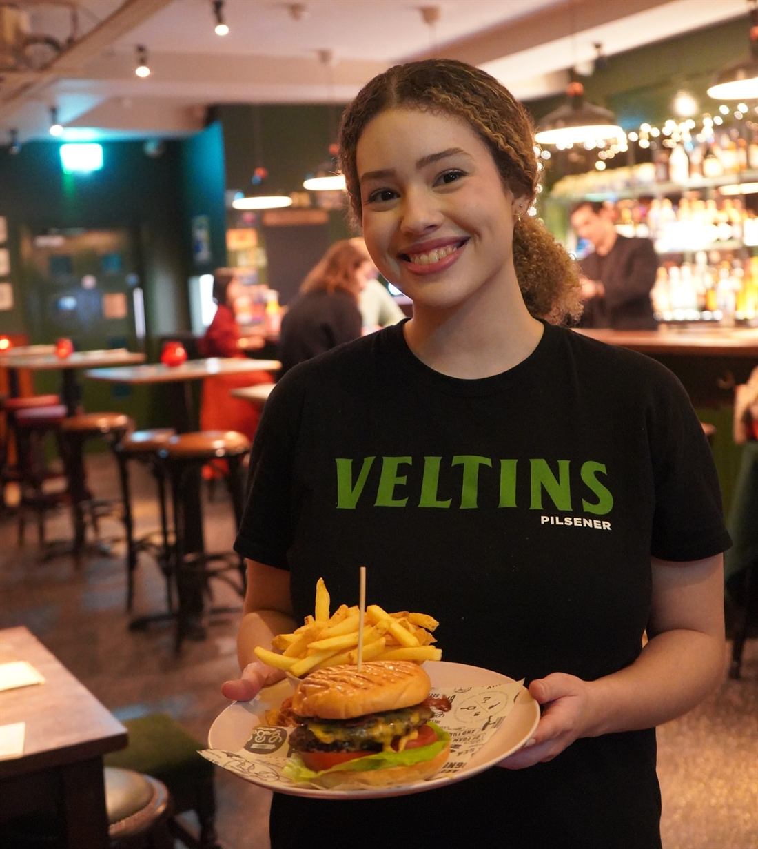 A smiling staff member carrying a stacked cheeseburger with fries to a table, the candlelit bar behind.