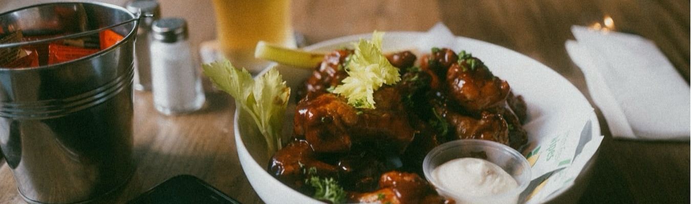 A bowl of sticky glazed wings with celery and a pot of dip beside a cold pint.